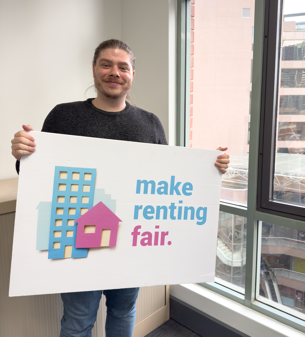 Dylan holding a sign saying make renting fair in front of a window.