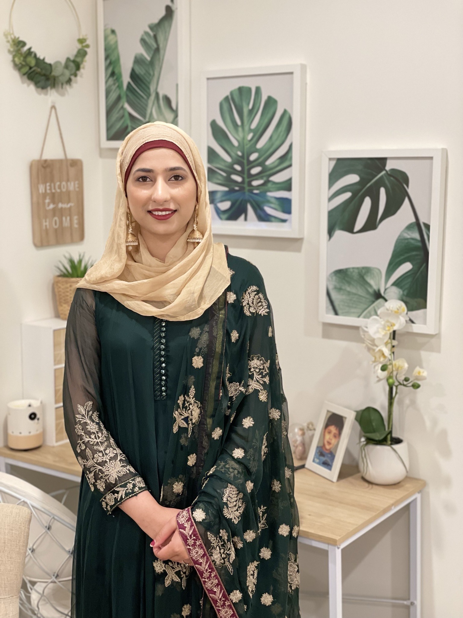 A woman smiles at the camera, standing in front of a desk and some artwork on the walls. She is wearing a dress and head scarf, with red lipstick and gold earrings.