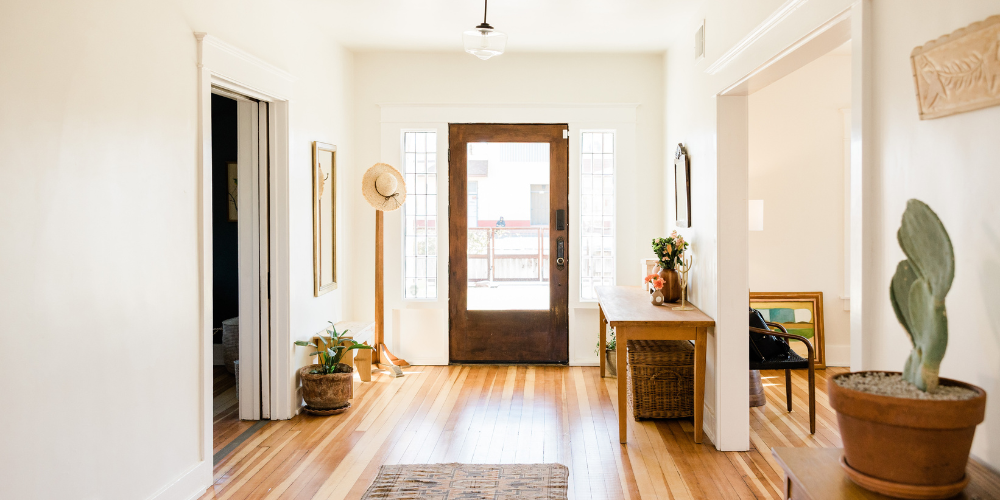 Entry way of a house. Brown front door, with a hat stand with a hat on it to the left. To the right, a table with flowers in a vase.