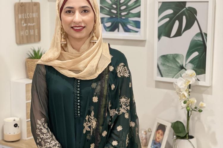 A woman smiles at the camera, standing in front of a desk and artwork on the walls. She is wearing a green dress, golden headscarf, with red lipstick and golden earrings.