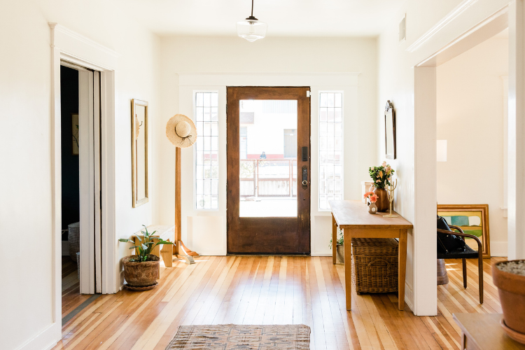 Entry way of a house. Brown front door, with a hat stand with a hat on it to the left. To the right, a table with flowers in a vase.