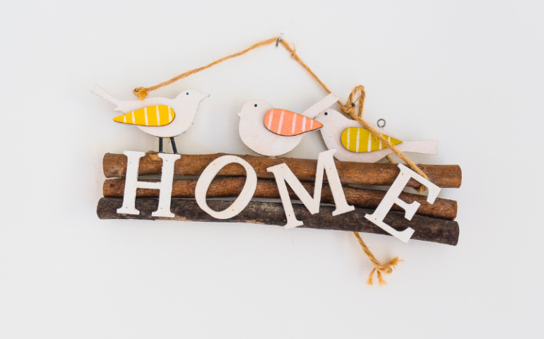 A dark wooden sign against a blank wall with the word home on it. Wooden birds sit on top.