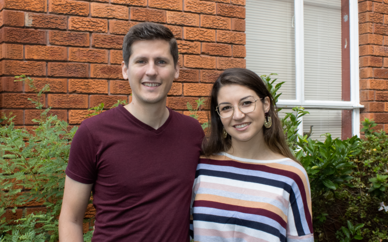 Jason and Lauren are standing close and smiling in front of a red brick wall and white window.