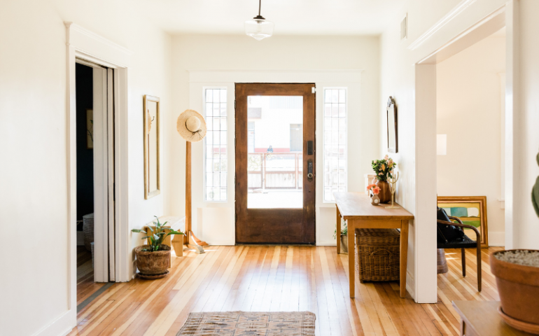 Entry way of a house. Brown front door, with a hat stand with a hat on it to the left. To the right, a table with flowers in a vase.